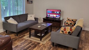 Una sala de estar con un sofá gris, un sofá de cuero marrón y un sillón gris. Una mesa de centro de madera está en el centro. Un televisor se encuentra sobre un mueble de madera a la derecha.