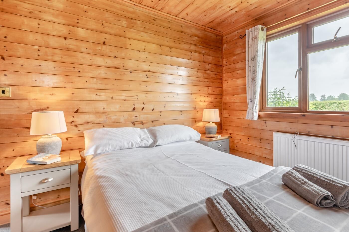 A wooden cabin room with a bed, two bedside tables with lamps, a window with curtains, and a radiator.