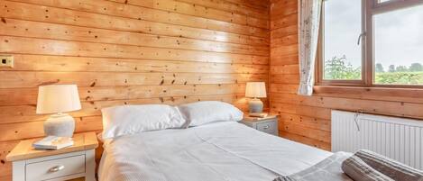 A wooden cabin room with a bed, two bedside tables with lamps, a window with curtains, and a radiator.