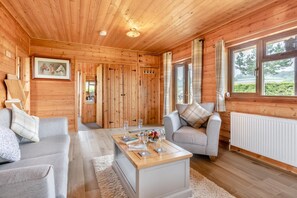 A log cabin interior with a sofa, a coffee table with glasses, a fireplace, and a view through the windows. - 2 Bed in Somerton (oc-w29203) (Somerton)