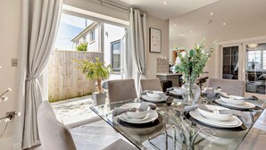 A dining area with a glass table set for six, surrounded by chairs, with a view of a wooden fence and a potted plant outside.