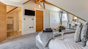 A bedroom with a sloped ceiling, a bed, a desk with a mirror, a TV, and a bottle of champagne.
