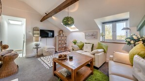 Una sala de estar tipo loft acogedora con techo alto, viga de madera, tragaluz, televisor de pantalla plana, sofá blanco, mesa de centro de madera y un ventanal con vistas.