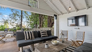 Un espace extérieur accueillant, avec une terrasse en bois, un canapé agrémenté de coussins à rayures, une table basse ronde avec des bougies et un téléviseur fixé au mur.