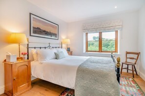 A neatly made bed with a headboard, two bedside lamps, a nightstand with a tissue box and a vase of flowers, a window with a view of greenery, and a wooden chair. - 3 Bed in Withypool (oc-o18550) (Withypool)