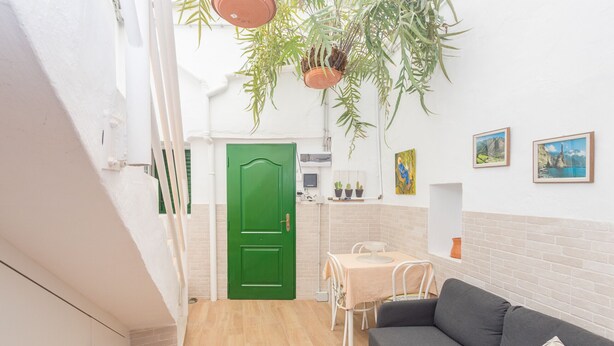 Un espacio habitable compacto con una puerta verde, una zona de comedor con mesa y sillas, un sofá y plantas colgantes.