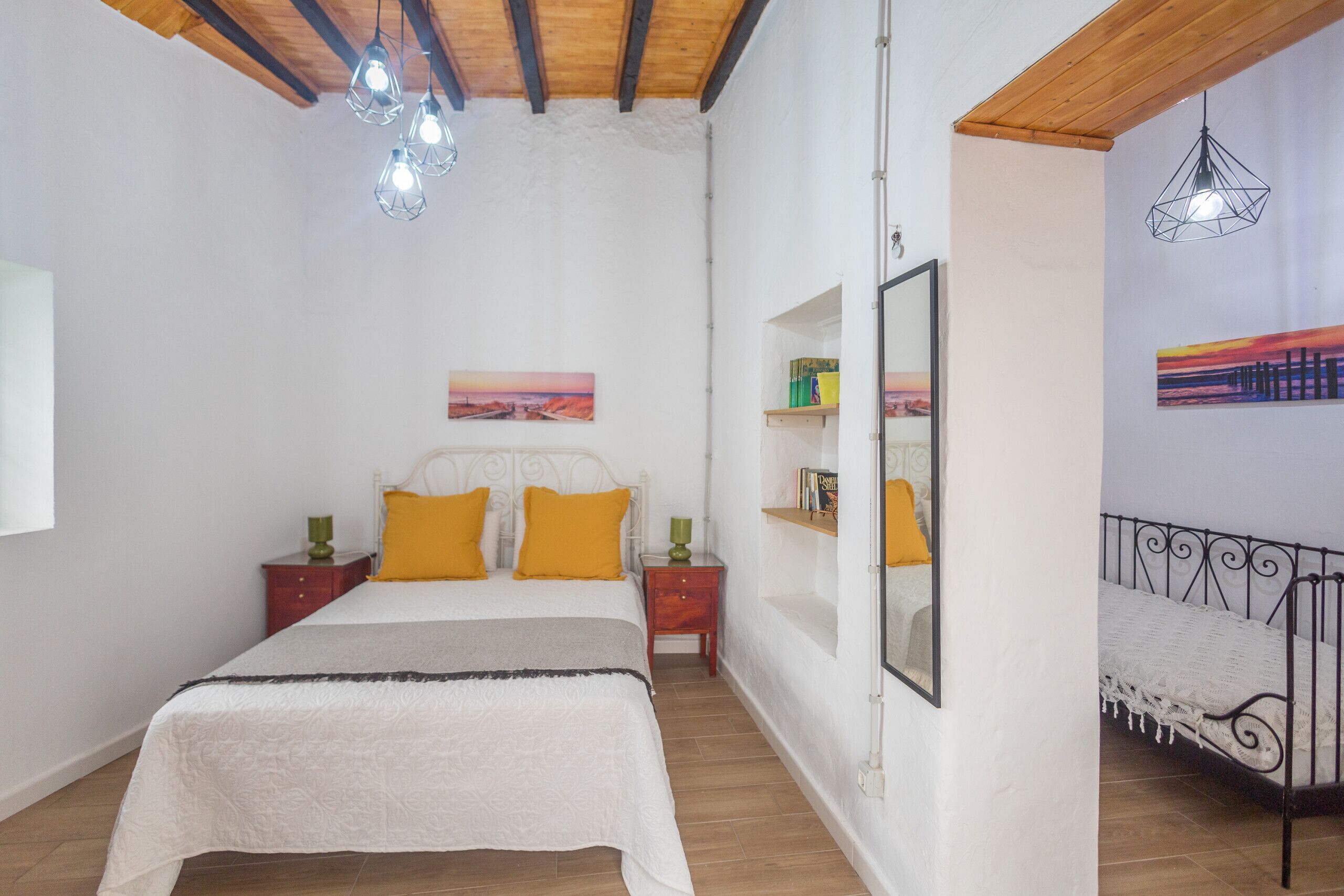 A bedroom with a bed, wooden ceiling, wall-mounted light fixtures, a bedside table, a mirror, and a shelf with books.