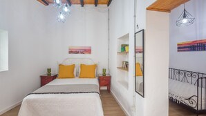 Un dormitorio con cama, techo de madera, lámparas fijadas a la pared, una mesita de noche, un espejo y una estantería con libros.
