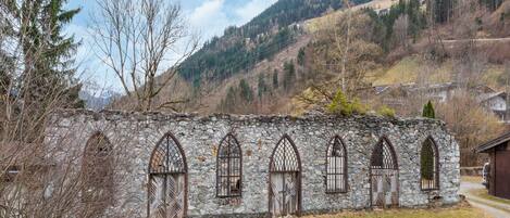 A stone building with arched windows and a stone wall, surrounded by trees and a mountainous backdrop.