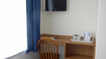Um quarto pequeno com uma cama, uma escrivaninha com cadeira, uma televisão e uma janela com cortinas.