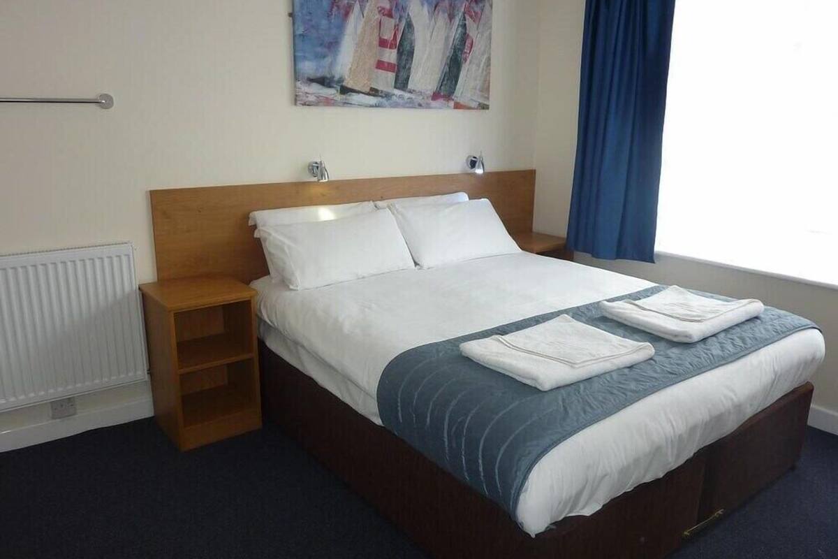 A neatly made bed with white and blue bedding, a wooden headboard, a bedside table, and a painting of sailboats on the wall.