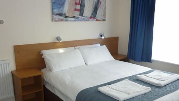 Habitación de hotel con cabecera de madera, cama con ropa blanca, manta azul y un cuadro con barcos a vela en la pared.