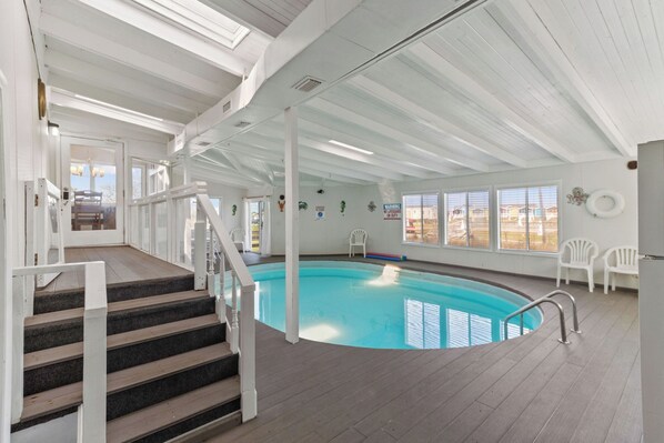 An indoor swimming pool with a wooden deck, white railings, and a view of the outside. - Isle Be Back Coastal Cottage, Private, heated, indoor pool (Hatteras)