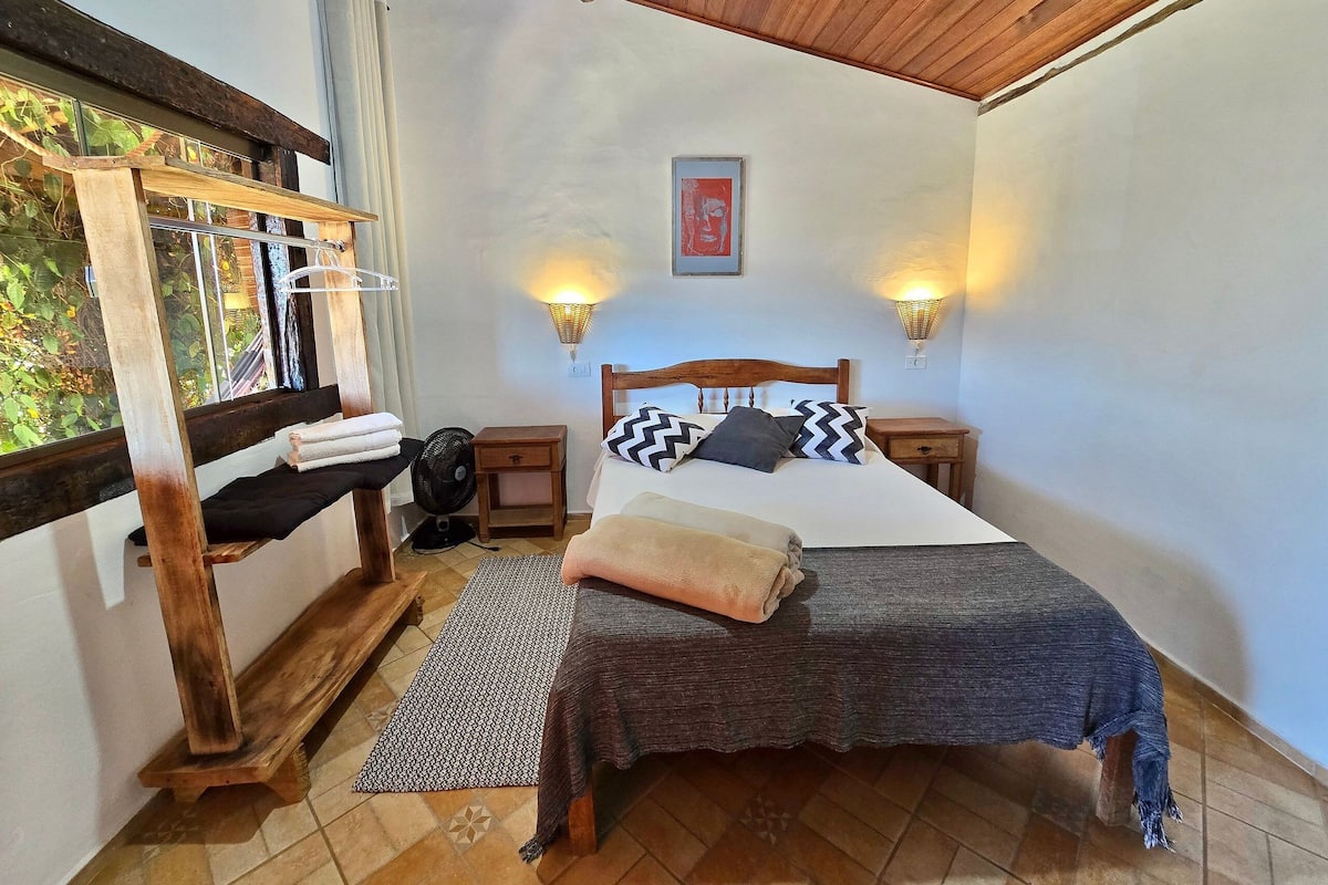 Un dormitorio con una cama de madera, dos mesitas de noche con lámparas, un banco y una ventana con cortinas.