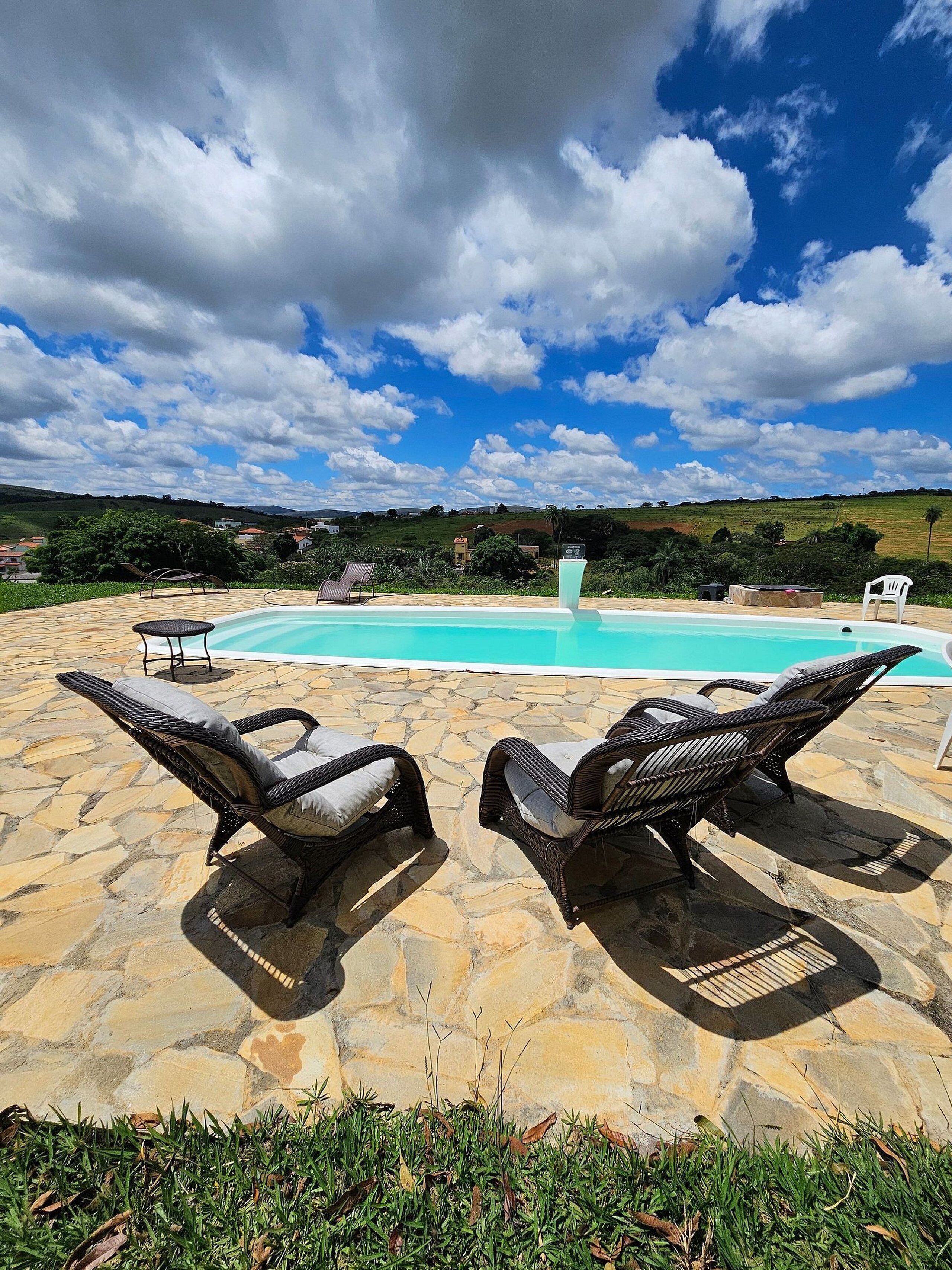 A pool area with stone flooring, wicker lounge chairs, and a view of a hilly landscape in the background.