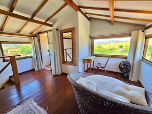 Un dormitorio con techo de madera, una cama de mimbre, una mesita, una silla y vistas a un paisaje verde a través de una ventana.