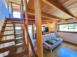 Un salón espacioso con techo alto, una escalera de madera, un sofá con cojines verdes y una cocina al fondo.