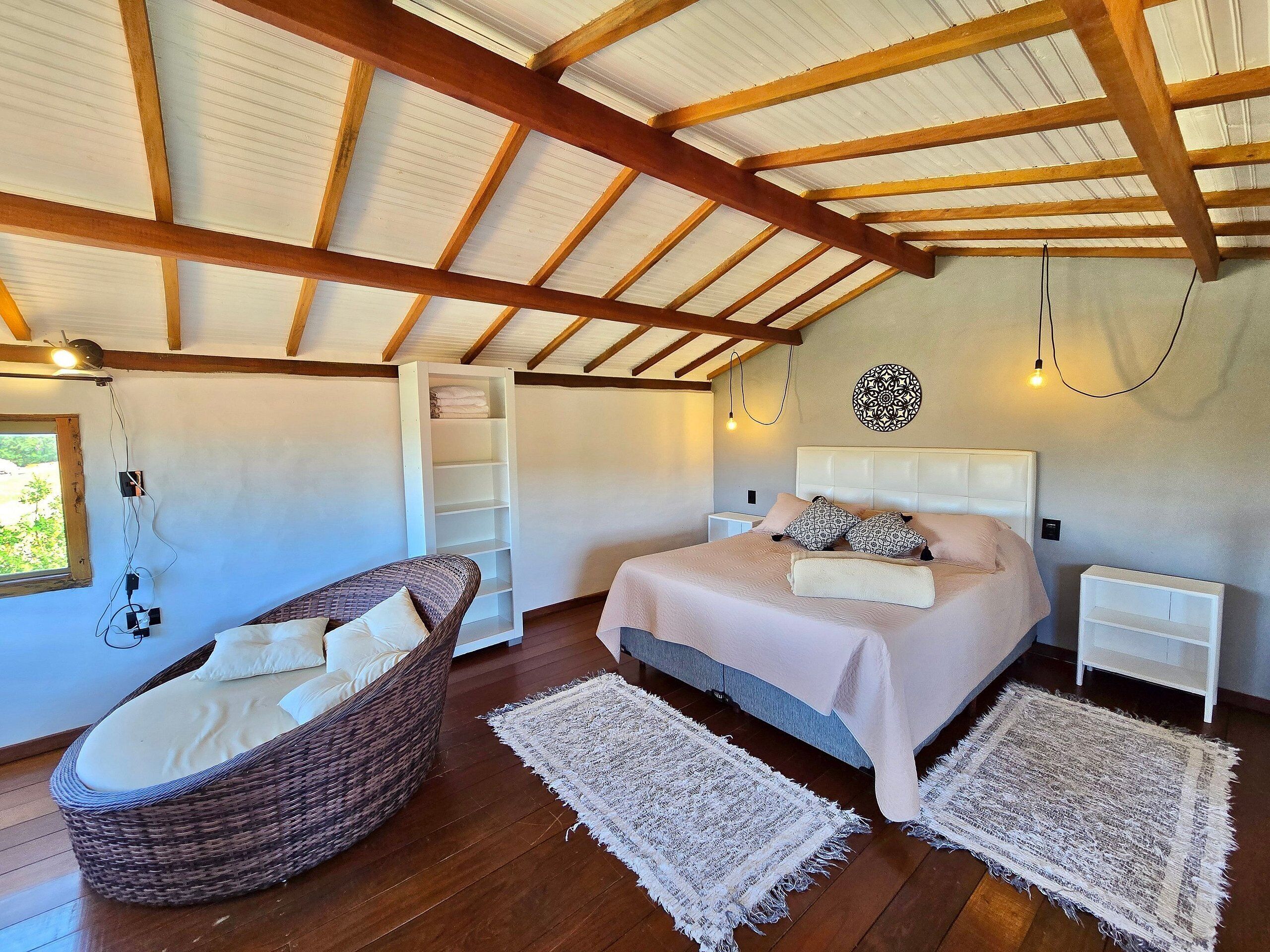 Un dormitorio con una cama, mesitas de noche, una silla de mimbre, un estante y una ventana.
