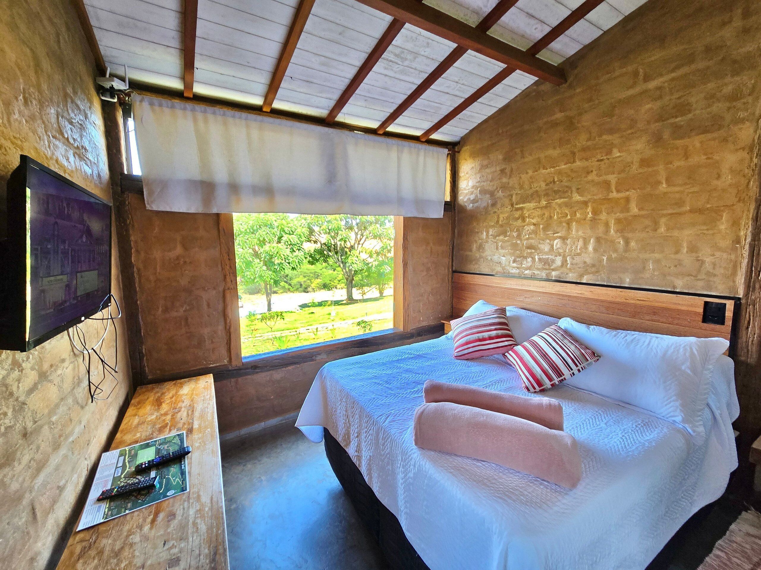 A wooden room with a bed, a TV mounted on the wall, and a window with a view of greenery.
