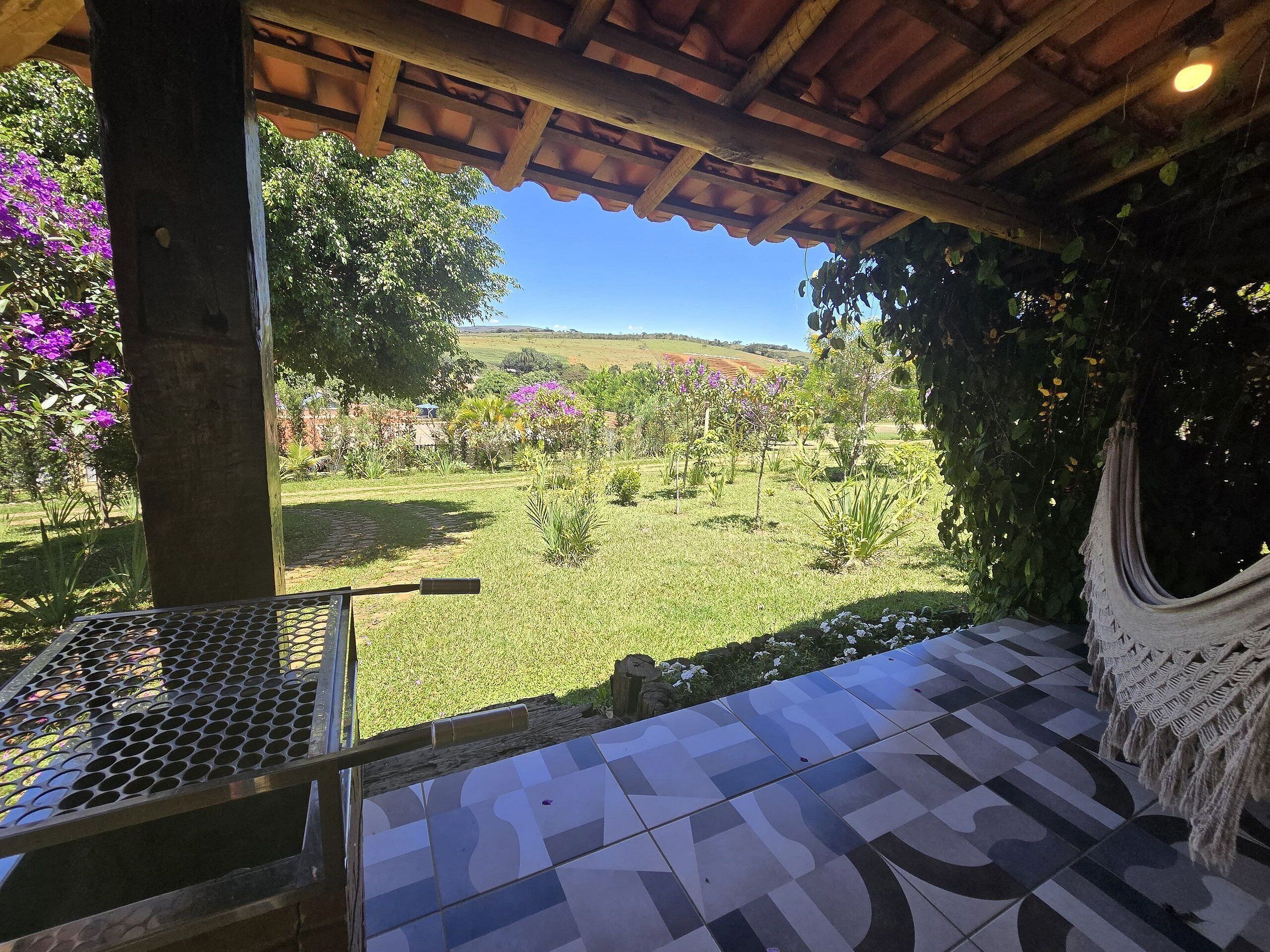 A covered patio with a patterned tile floor, a hammock, and a view of a landscaped garden with various plants and a distant hill.