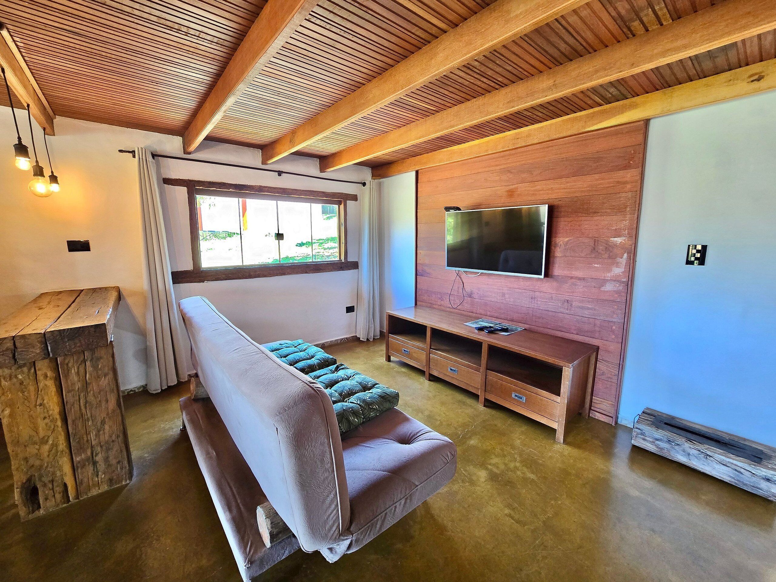 A cozy room with a wooden ceiling, a large window, a flat-screen TV mounted on a wooden wall, a bench, and a small wooden table.