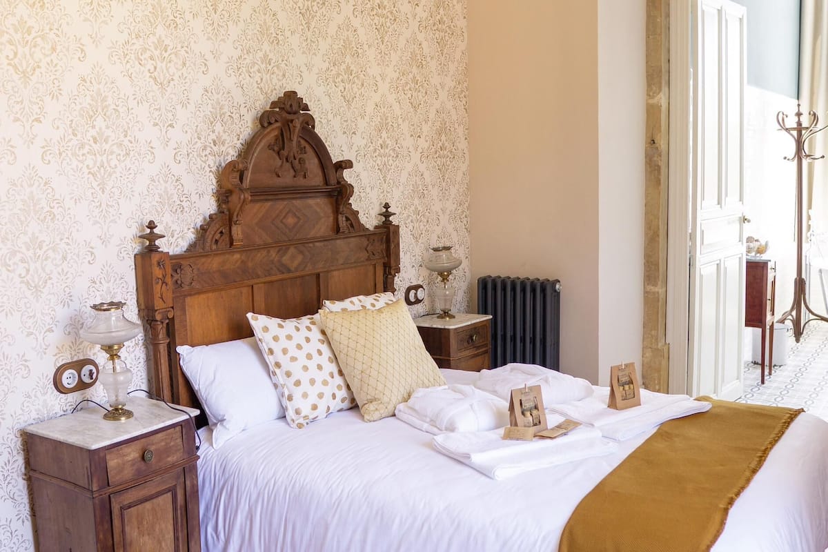 Un dormitorio con cabecera de madera, cama con ropa blanca, mesita de noche con lámpara y radiador.
