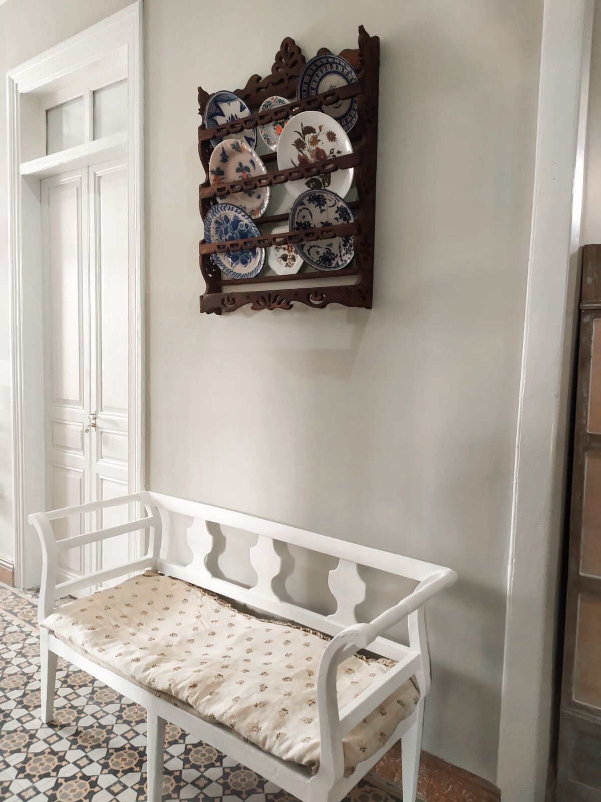 Un banc blanc avec un coussin à motifs, dans une pièce dont le sol est carrelé et où est fixée au mur une étagère à assiettes.