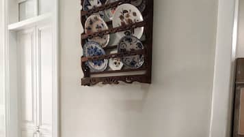 Un banc blanc avec un coussin à motifs, dans une pièce dont le sol est carrelé et où est fixée au mur une étagère à assiettes.