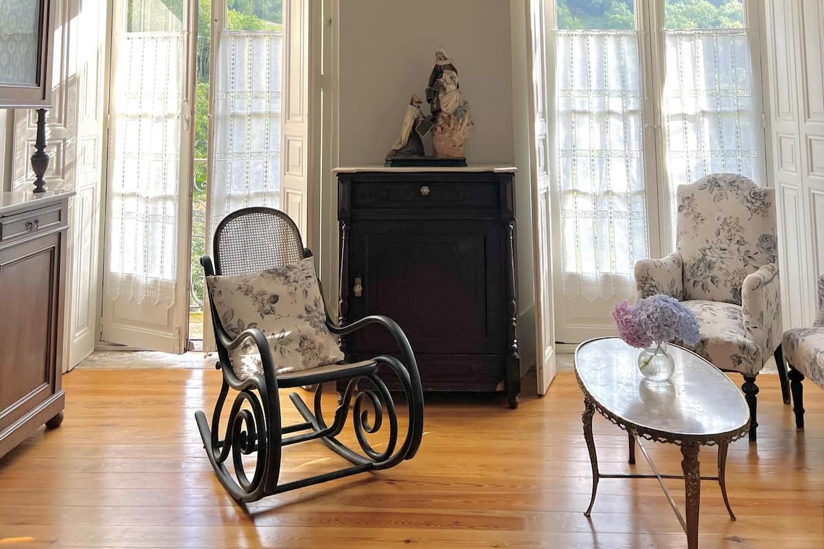 Una habitación con candelabro, dos ventanas grandes con cortinas, chimenea, sillón mecedora y una mesa con flores.