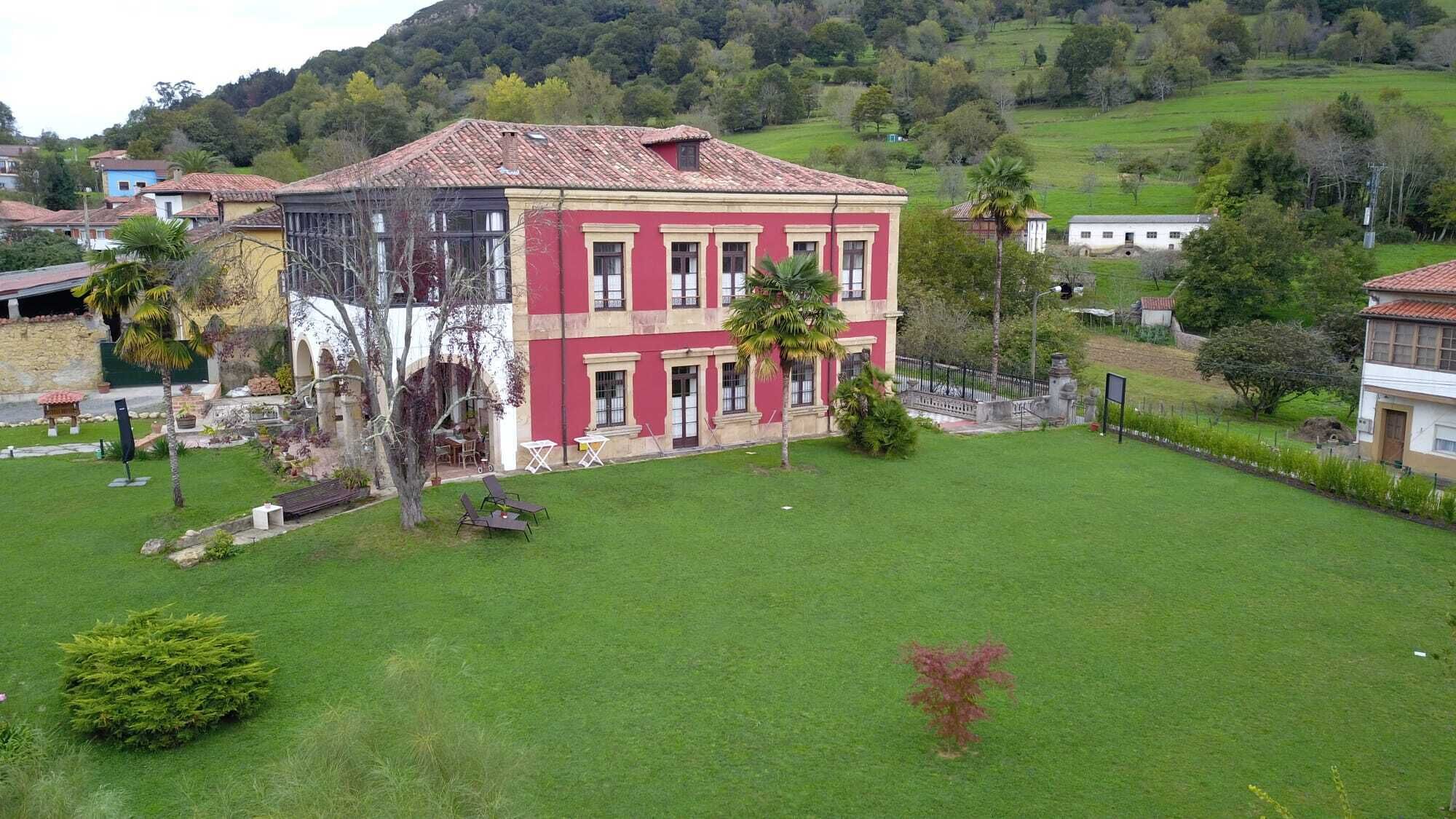 Un edificio de dos pisos con fachada roja y techo de tejas, rodeado de un césped cuidado y algunos árboles.