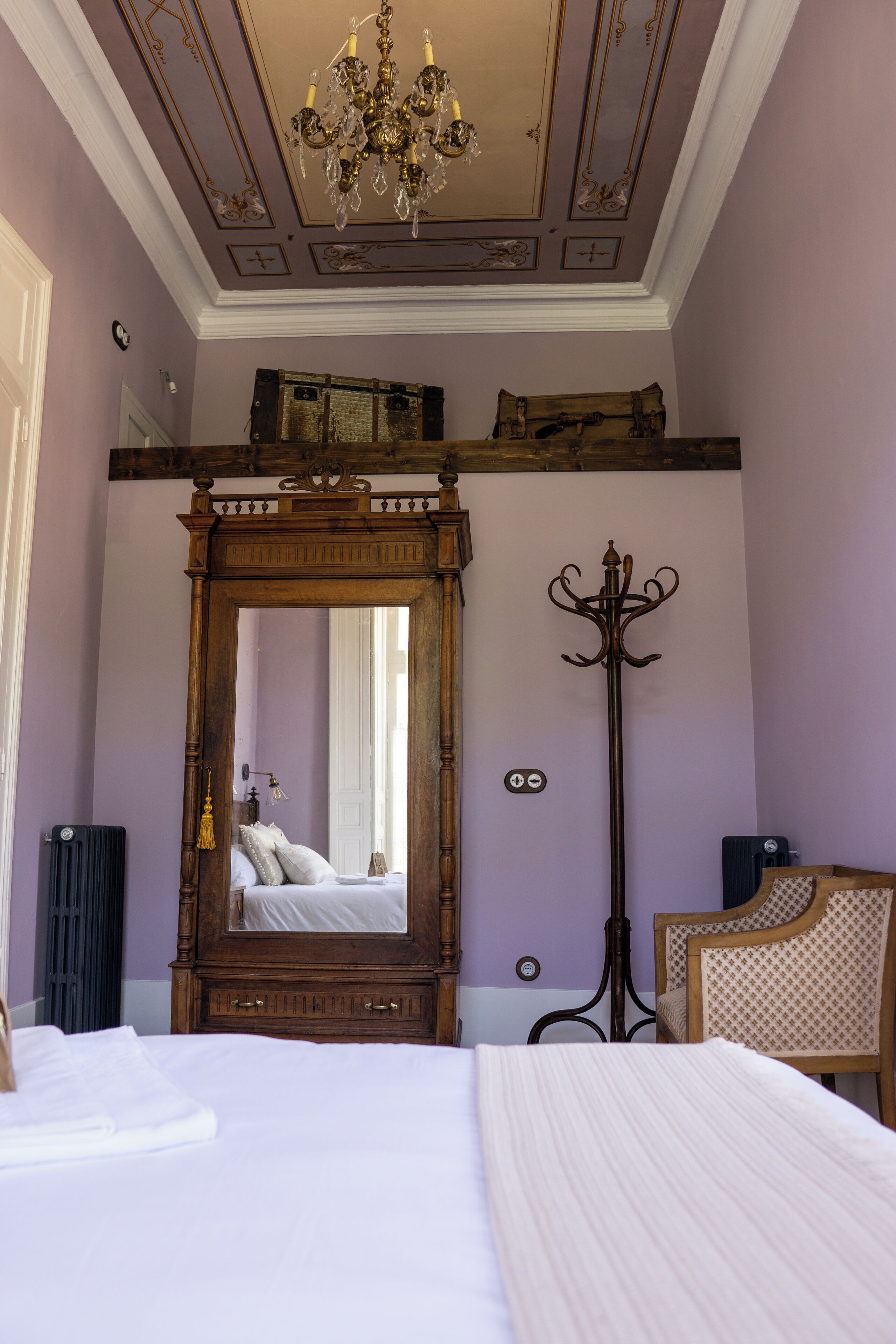 A bedroom with a large mirror, a bed with white linens, a wooden dresser, and a chandelier.