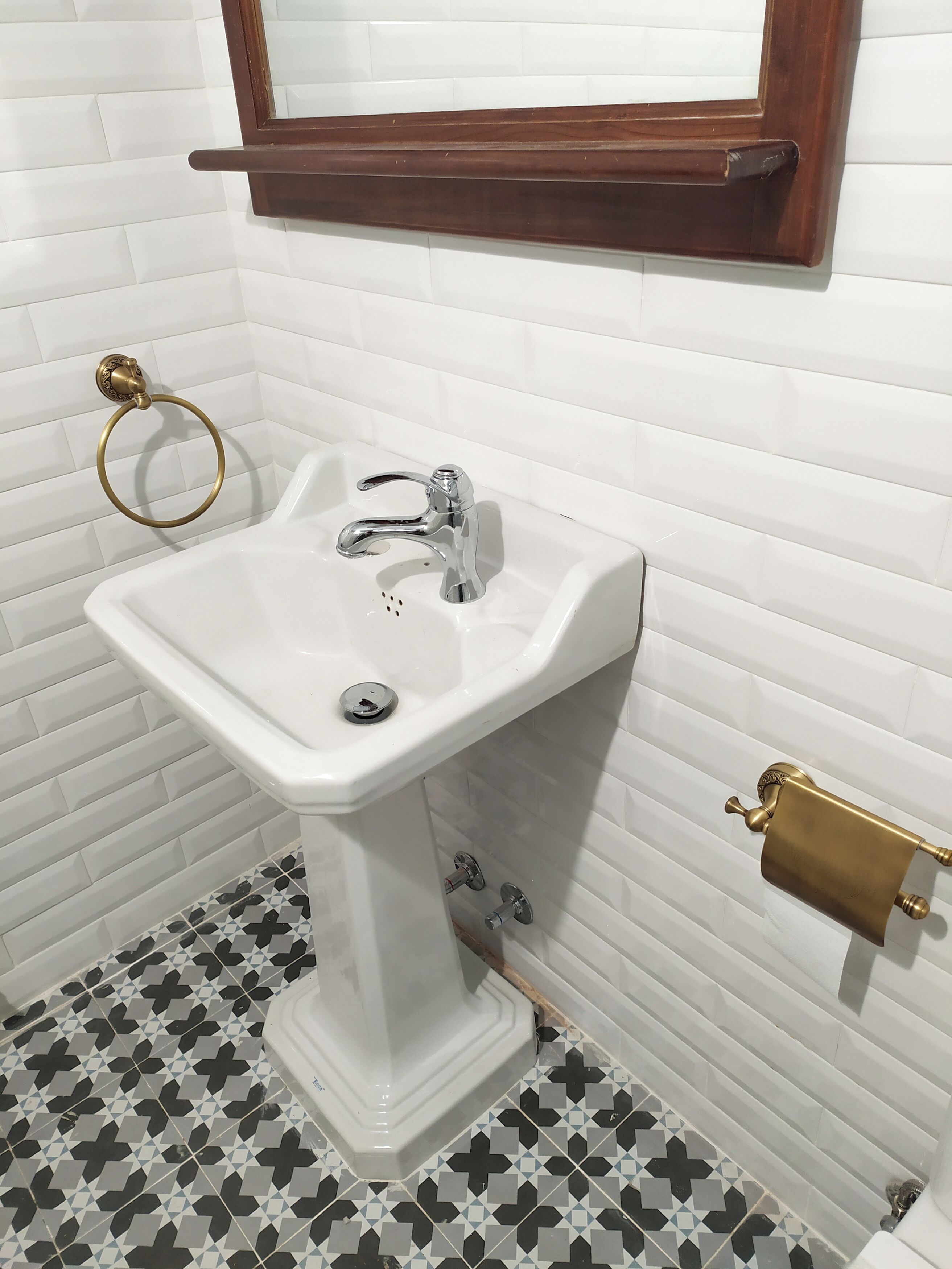 A bathroom with a white pedestal sink, a gold towel ring, a wooden towel rack, and a toilet paper holder. The floor has a black and white geometric tile pattern.