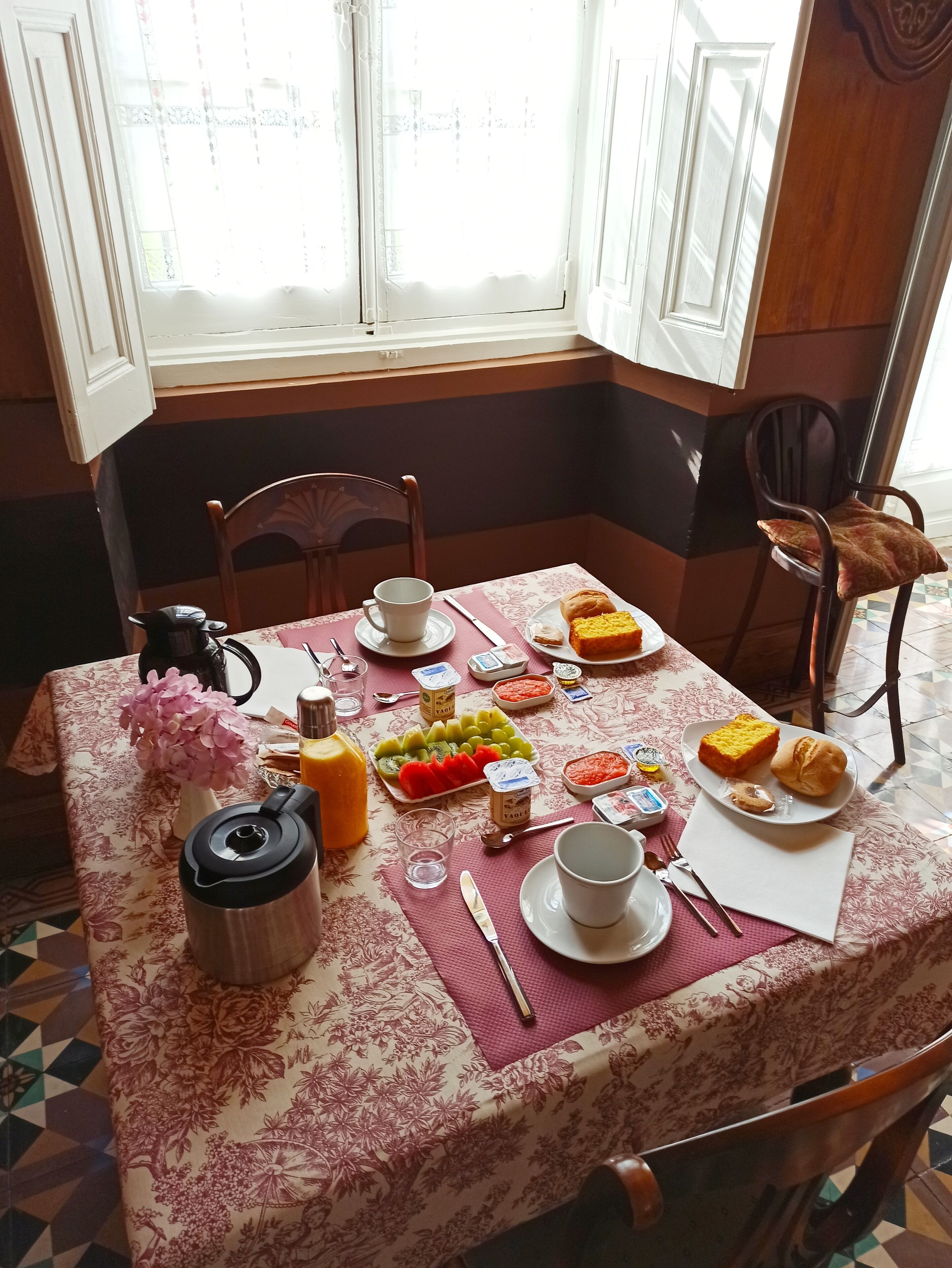 Taula de menjador coberta amb un llençol floral, diversos plats incloent fruita, pa i condiments, i una cafetera.