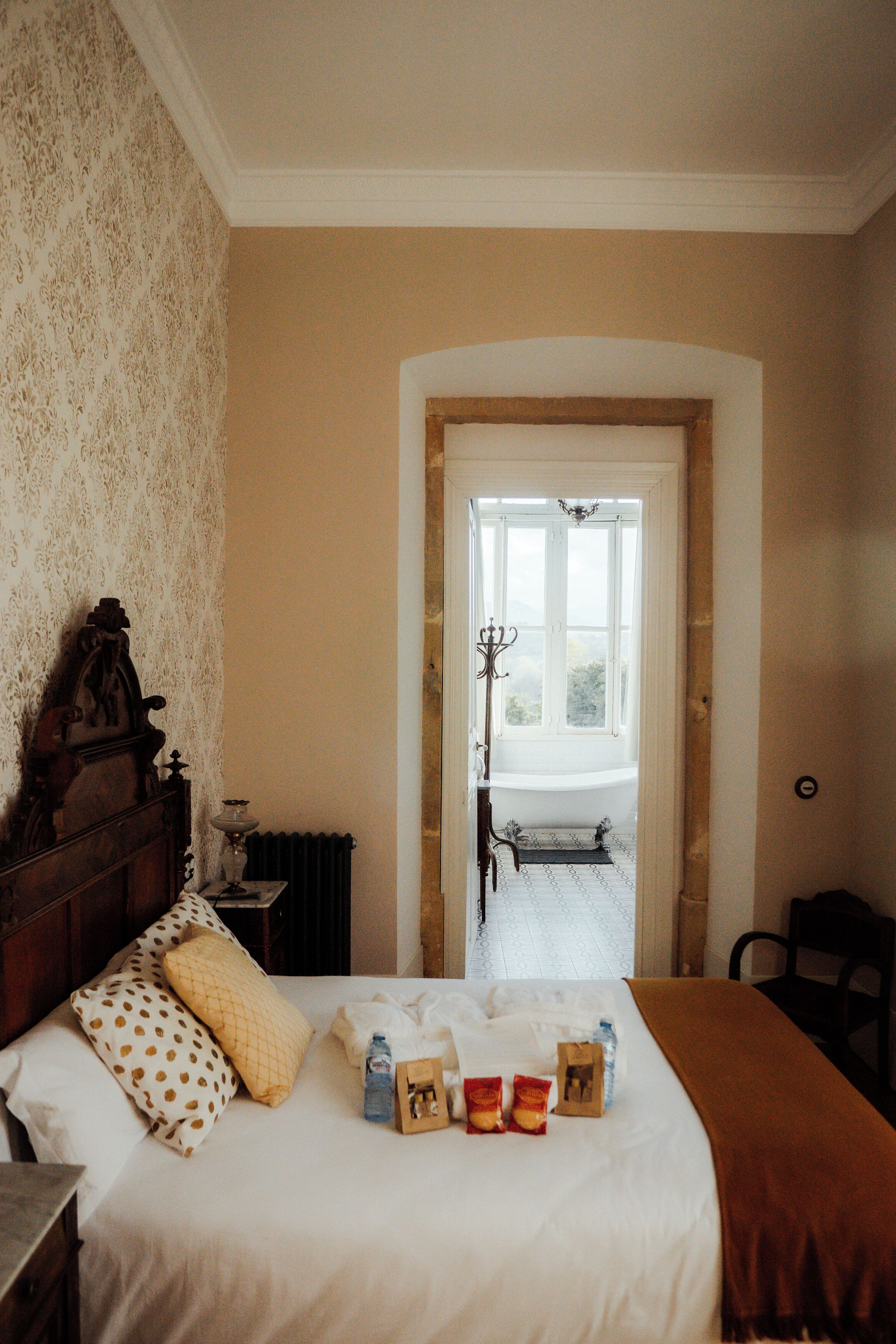 A hotel room with a bed, a wooden headboard, a radiator, and a view of a balcony through an arched doorway.