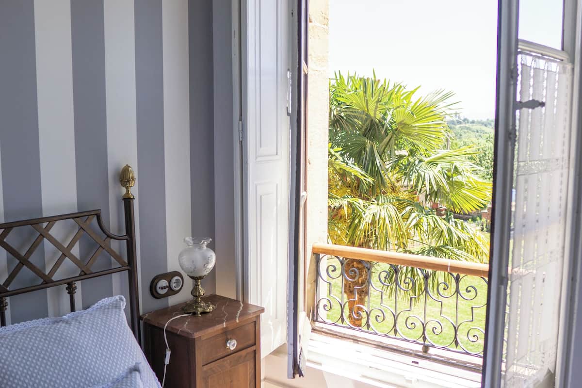 Un dormitorio con cama, mesita de noche, lámpara y vistas a un balcón con plantas.