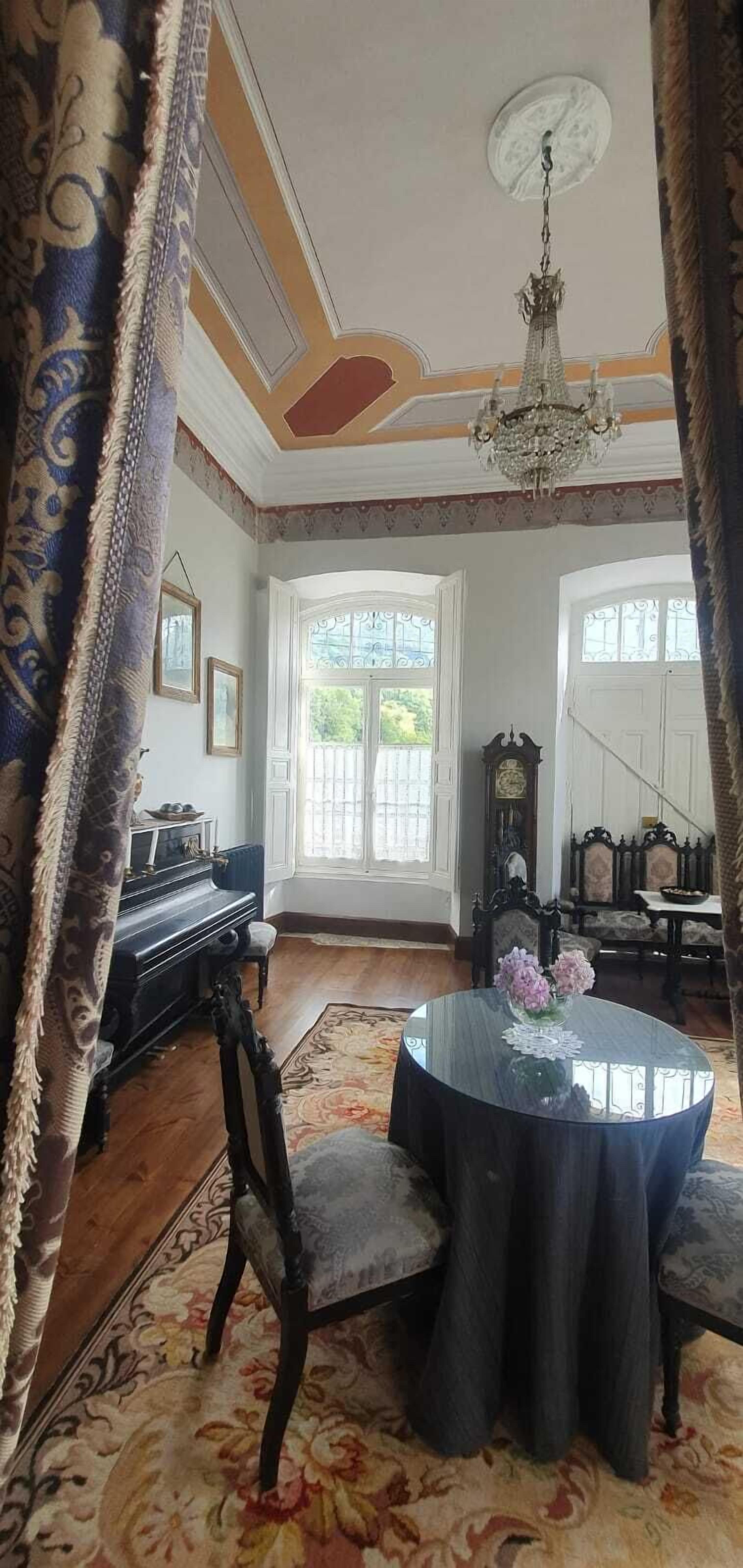 A room with a piano, a round table with a vase of flowers, and a chandelier.