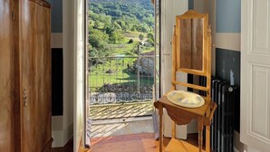 Een kamer met houten vloer, een houten stoel, een witte deur die leidt naar een balkon met uitzicht op bergen, en een radiator.