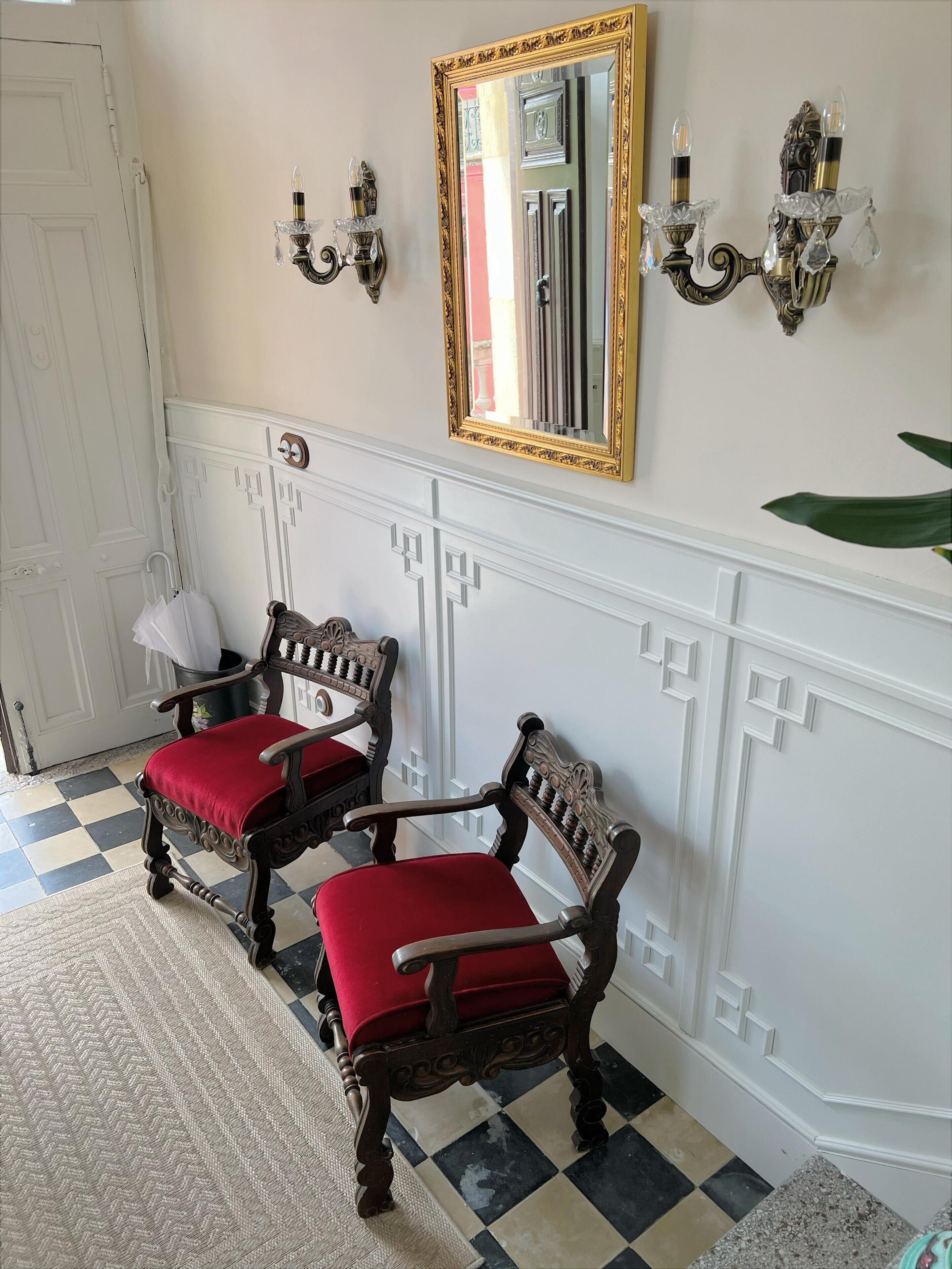 Ein Zimmer mit zwei verzierten Holzsesseln mit roten Kissen, einem großen Spiegel mit goldener Rahmung und Wandleuchten mit Kristallverzierungen.