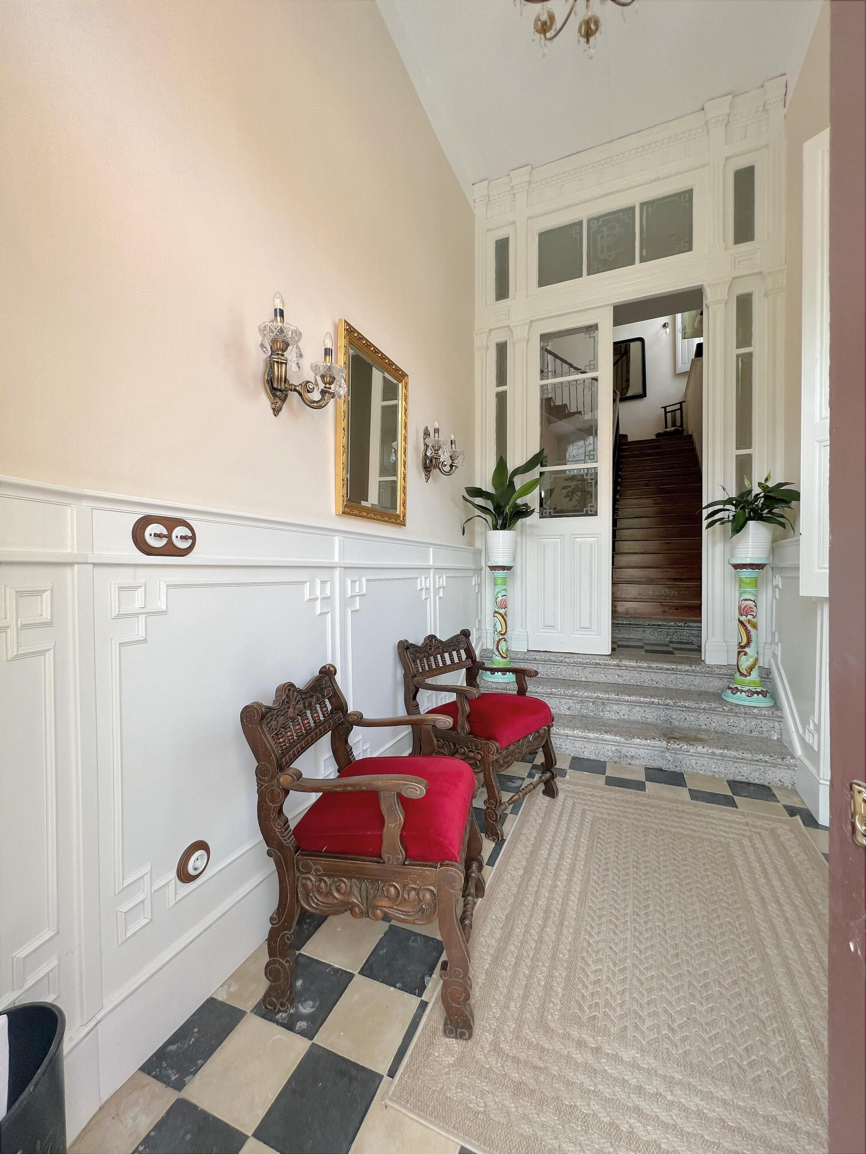 Un couloir avec deux fauteuils en bois richement décorés, dotés de coussins rouges, un sol à carreaux et un escalier menant à un étage supérieur.