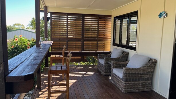 Ein überdachter Patio-Bereich mit Holztisch und -stühlen, einer Rattan-Couch und einem Fenster mit Jalousien.