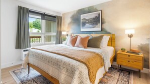 A bedroom with a large bed, a nightstand with a lamp, a painting on the wall, and a window with curtains. - Cityside Escape (Branson)