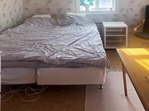 Una cama con cubre blanco, una mesita de noche, una silla y una ventana con una planta.