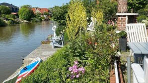 A riverside garden with a boat docked, a brick chimney, and a white chair. - Cosy Holiday Home Near the Centre (Krummhoern)