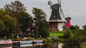 A traditional windmill with a red and white structure, a grassy area with a small bridge, and boats on a calm water body. - Cosy Holiday Home Near the Centre (Krummhoern)