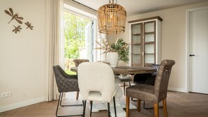 A dining area with a wooden table, four chairs, a hanging light fixture, and a view of greenery outside.