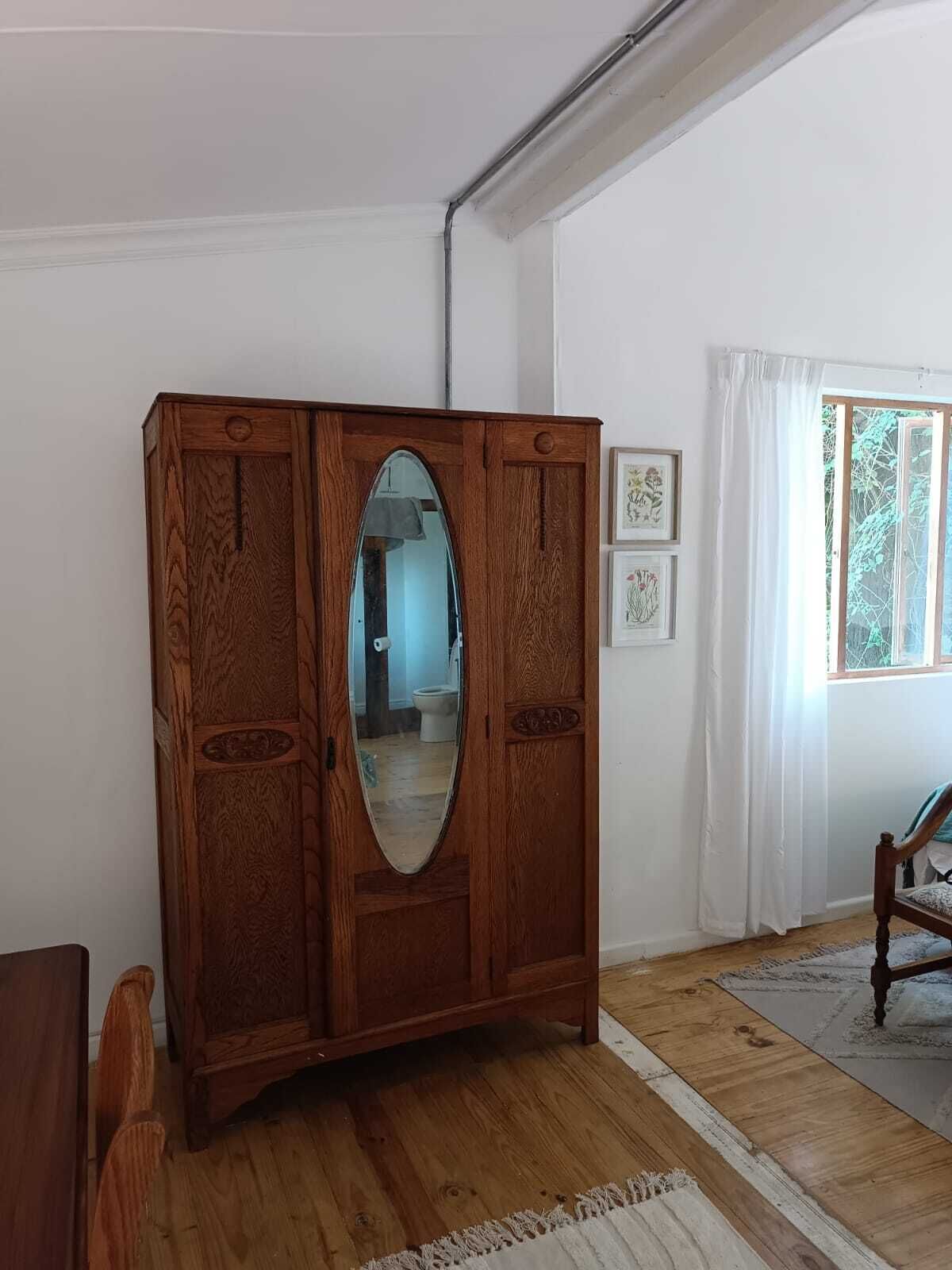 A wooden armoire with an oval mirror, a wooden chair, and a window with white curtains.