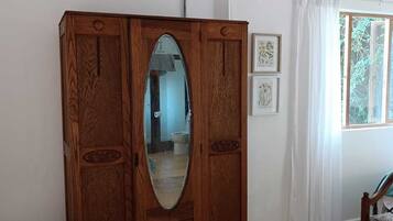 A wooden armoire with an oval mirror, a wooden chair, and a window with white curtains.