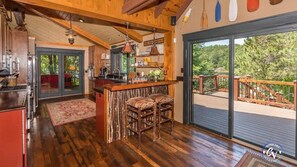 Une cuisine rustique avec des poutres en bois, un coin bar avec deux tabourets et une grande porte-fenêtre coulissante donnant sur une terrasse extérieure.