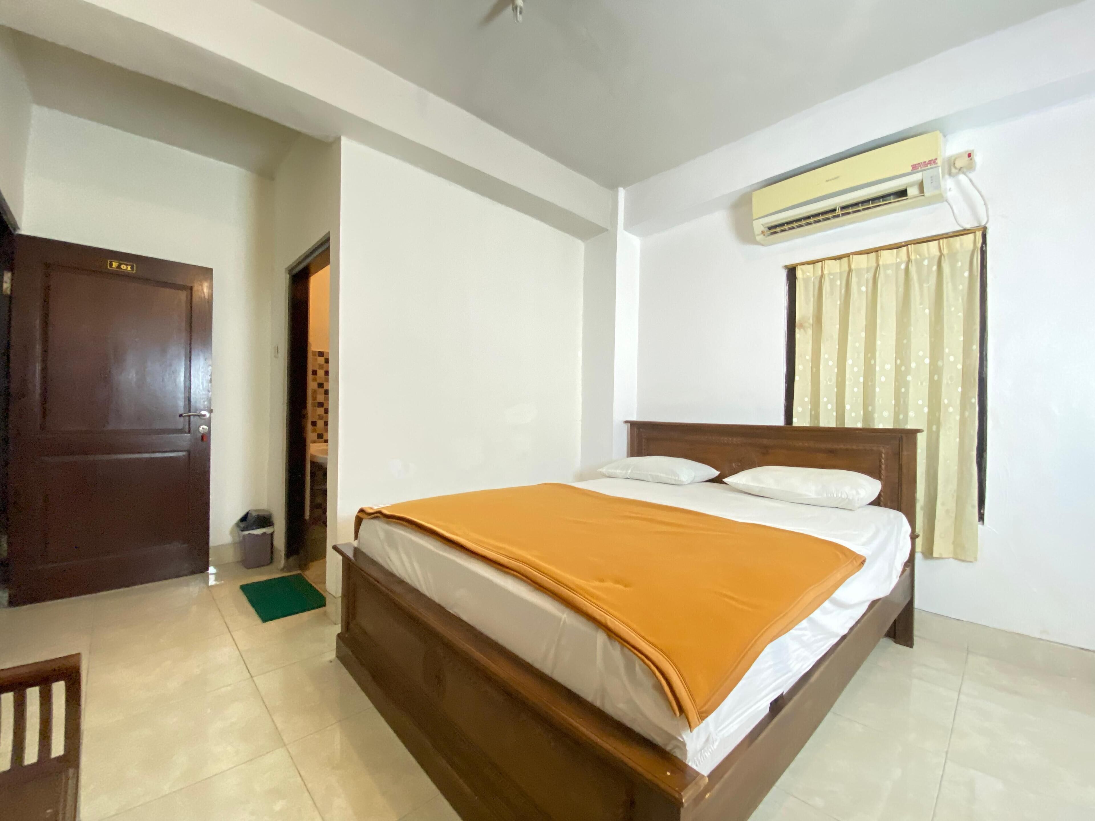 Une chambre d’hôtel avec un lit en bois, des murs blancs, un tapis vert, une porte et un appareil de climatisation.