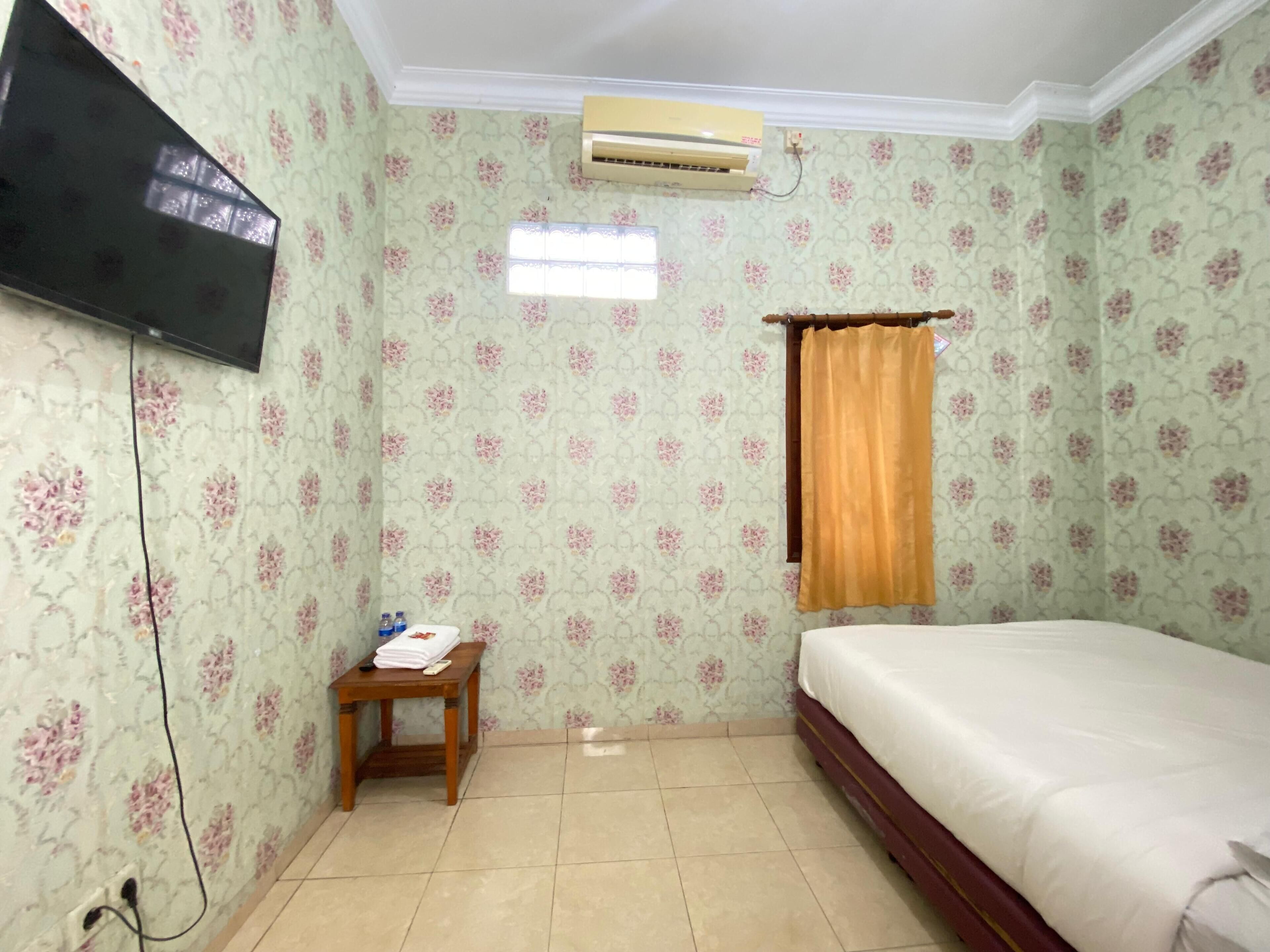 A bedroom with floral wallpaper, a bed, a TV, an air conditioner, a small table with items, and a window with a curtain.